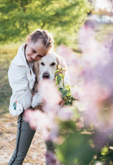 Selective focus. Close-up portrait of a beautiful girl with two pigtails in white jacket. She is hugging her golden retriever and smiling. Lilac bush on the foreground. Sunset light.