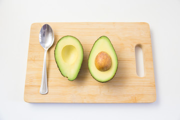 Spoon and halved avocado with core on wooden chopping board. Top view. Isolated objects on white background. Fresh food or healthy diet concept