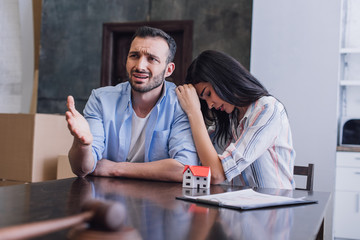 Selective focus of upset woman leaning to worried man at table with house model, documents and gavel in room - obrazy, fototapety, plakaty