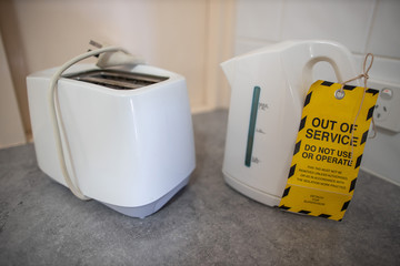 Safe workplaces yellow out of service tag placing on electricity kettle unit on the bench as faulty...