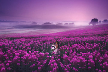 A beautiful girl in a pink dress standing with a lantern full of magic lights in a large pink field of willow herb. Romantic evening photo with sunset sky.
