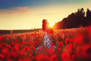 A beautiful young girl in a long dress stands in a poppy field. Silhouette sunset photo.