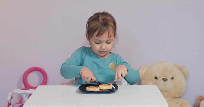 Cute little girl child with bright eyes eats curd fritters at home with spoon and knife. baby etiquette. Table manners