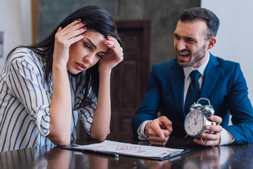 Angry collector holding alarm clock and pointing at documents near stressed woman at table in room