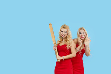Bad girls. Young blond women  in fashionable red dresses holds baseball bat or racket on blue background, copy space.