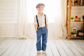 Cute boy in a white shirt, cap and suspenders in a bright room. Retro style in clothes - 348532559