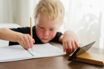 Little sad boy learning at home. Watching webinar by mobile, writing