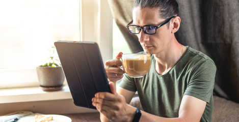 Young handsome man reading interesting news on the Internet using a gadget tablet sitting at a table near the window