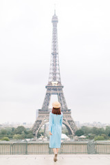 Back view of stylish tourist woman in blue dress and hat sightseeing Eiffel Tower in Paris, France