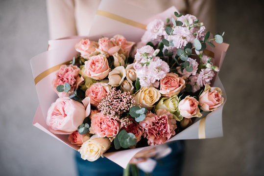 Very nice young woman holding big beautiful blossoming bouquet of fresh roses, matthiola, carnations, ozothamnus, eucalyptus flowers in tender pink colors on the grey wall background