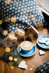 Fototapeta na wymiar Delicious fresh cappuccino coffee in a blue cup with some heart shaped festive cookies, fireflies, spruce leaves and christmas decorations (little wooden houses) on the wooden table background