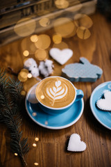 Delicious fresh festive blue cup of cappuccino coffee with spruce leaves, cotton, fireflies and festive cookies on the rustic wooden table background