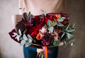 Very nice young woman holding big beautiful blossoming bouquet of fresh roses, carnations, berries, eucalyptus flowers in red, pink and burgundy colors on the grey wall background