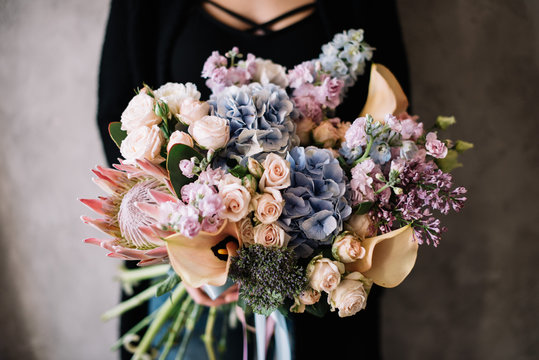 Very nice young woman holding big beautiful blossoming bouquet of fresh hydrangea, nutan protea, lilac, matthiola, roses, calla lilies flowers in blue, pink, cream and lilac colors 