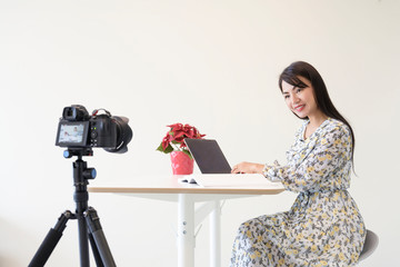 Beautiful asian woman sitting in room recording video online for social media, with latop on table smiling to camera on wall background.