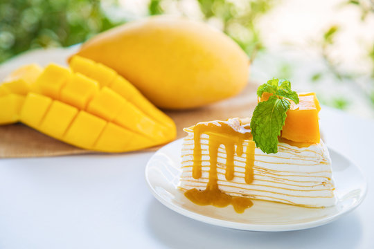 Mango crepe cake in white dish with peppermint and whip cream topping on white table with mango fruit and sliced background.