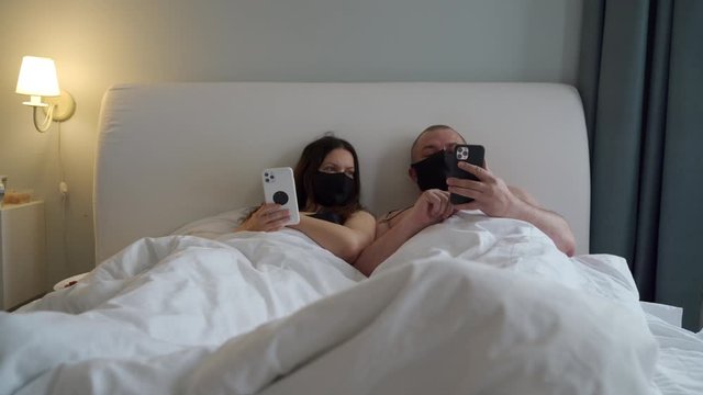 Adult married couple in protective medical face masks lying in bed during coronavirus COVID-19 quarantine at home, man and woman holding mobile phones.