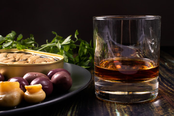 A glass of whiskey and an elite appetizer on a black background. Olives, pickled mushrooms, champignons, sprats and an expensive alcoholic drink.