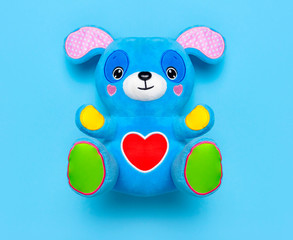 Blue soft children's toy puppy with funny ears, multi-colored paws and red heart on blue background flat lay top view copy space. Plush toy dog, baby friend, children's play item, happy childhood