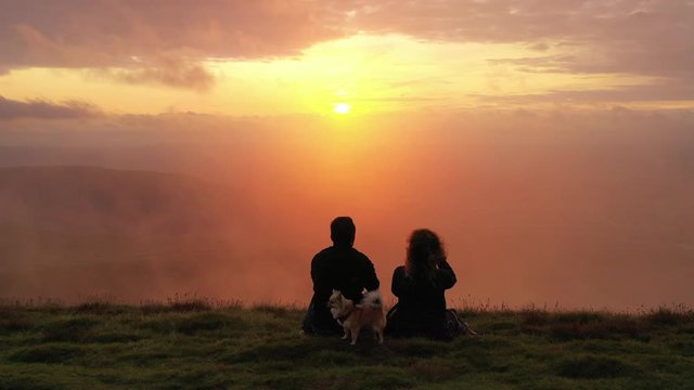 Silhouette couple sat watching beautiful sunset up scenic Welsh mountain view with cute chihuahua through wall of mist and fog