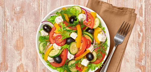 Greek salad with cucumeber, olives, feta cheese, cherry tomatoes, bell pepper and lettuce. Summer diet salad concept. Tasty greek salad in bowl on wood, top view