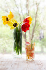 Glass of lemonade with grapefruit, orange, cucumber, ice  on wooden background with yellow and red tulips in vase on window
