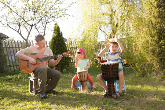 Happy Family Dad And Two Kids Having Fun With Musical Instruments Together Outdoors. Dad Playing  Guitar And Kids Playing Ethnic Drums. Quarantine. Musical Concept