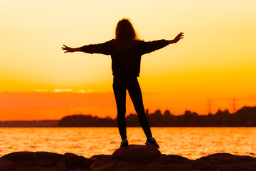 Woman's silhuette at sunset standing on rock looking straight with stretched arms. Nature and beauty concept. Orange sundown.