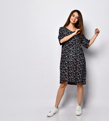 Brunette model in black dress with floral print, white sneakers. Smiling, touching her face, posing isolated on white. Full length