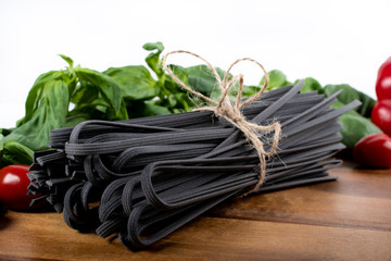 Black squid ink tinted tagliatelle pasta, uncooked, with basil and tomatoes in the background, on a wooden board