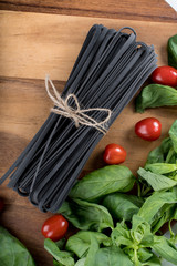 Black squid ink tinted tagliatelle pasta, uncooked, with basil and tomatoes in the background, on a wooden board