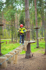 Boy climbing in an adventure park. In a green jacket and a red helmet. Overcomes obstacles.