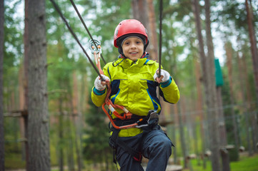 Boy climbing in an adventure park. In a green jacket and a red helmet. Overcomes obstacles.