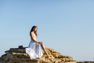 Happy brunette woman in a white dress sitting on a rock on a background of blue sky.