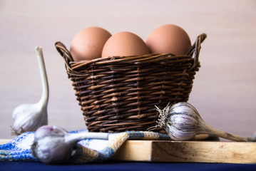Chicken eggs. Brown chicken eggs in basket on the cutting board on blue background. Copy space. Still life with eggs, garlic, onion. Farm natural products. Egg and vegetables is ingredient for cooking