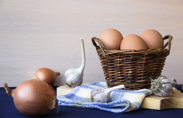 Chicken eggs. Brown chicken eggs in basket on the cutting board on blue background. Copy space. Still life with eggs, garlic, onion. Farm natural products. Egg and vegetables is ingredient for cooking