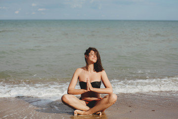 Fototapeta na wymiar Young beautiful woman practicing yoga on the beach, sitting on sand and meditaning. Mental health and self care concept. Happy girl relaxing on seashore on summer vacation