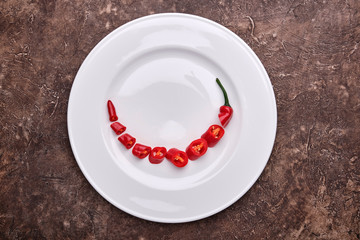 Red, searing, chilli pepper cut into pieces lies on a white plate on a brown background. View from the top.
