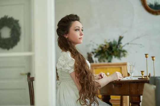 Beautiful woman with long hair in a long white dress. He sits at the table and looks away. Historical reconstruction