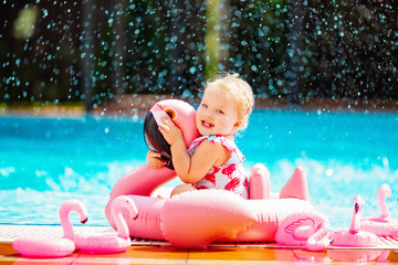 Baby blonde girl sitting in water spray with rubber pink flamingo near the pool