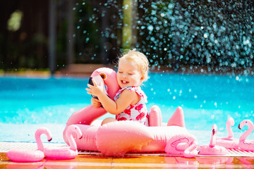 Baby blonde girl sitting in water spray with rubber pink flamingo near the pool