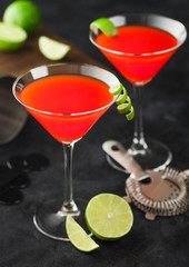 Cosmopolitan cocktail in classic crystal glasses with lime peel and fresh limes with strainer on black background.