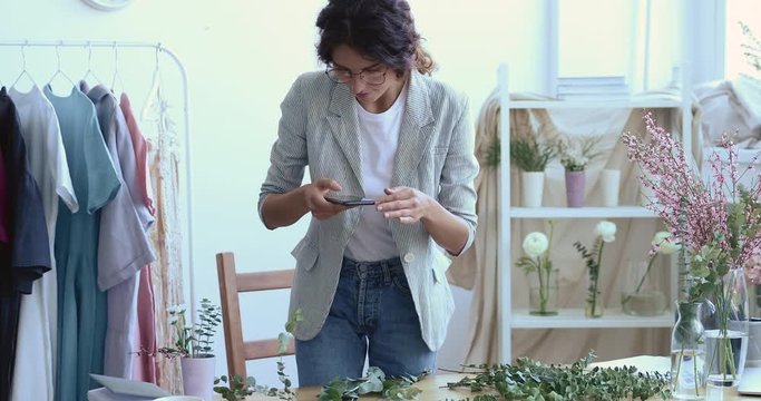 Young female designer florist blogger taking picture of floral plants decoration composition on smart phone working in modern creative studio. Handmade creative hobbies social media blogging concept.