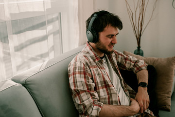 Young hipster millennial man sits on the couch and listens to music in wireless headphones. Modern lifestyle technology. Using phone. Connection to internet. Read message and answer phone calls.