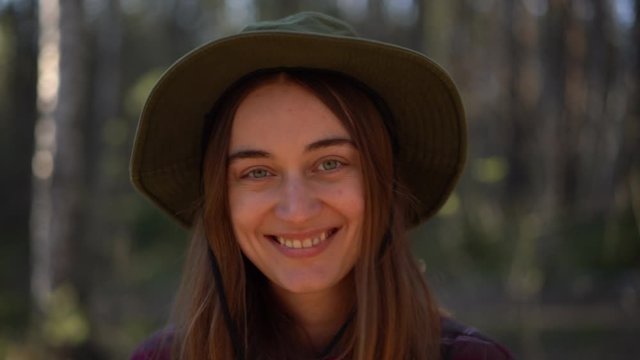close-up portrait joyful young caucasian hipster female in forest smiling looking at camera. Beautiful millennial woman in hat, hiking equipment enjoy camping day  nature, adventure freedom concept