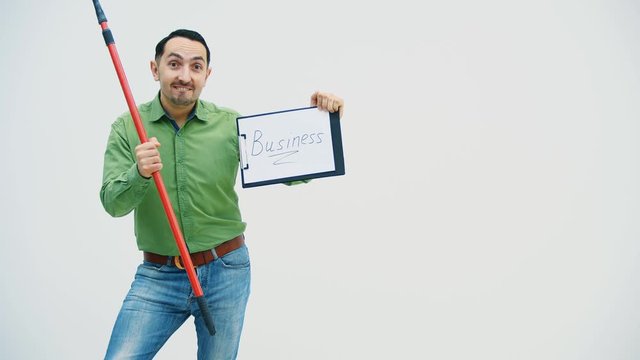 Funny young man standing with the paper holder with word "bisiness" writen on it, looking silly, holding and moving a broomstick.