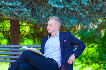 Handsome businessman in blue suit is sitting on the bench in summer park reading a book having rest after a hard working day