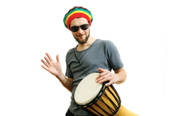 Young caucasian man in rasta hat, sunglasses and grey t-shirt on white background play on djembe...