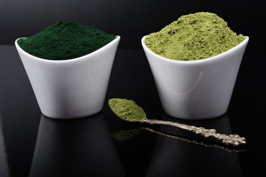 An isolated tablespoon of dried organic wheat grass and spirulina powder, on white rustic background