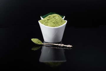 An isolated tablespoon of dried organic wheat grass and spirulina powder, on white rustic background